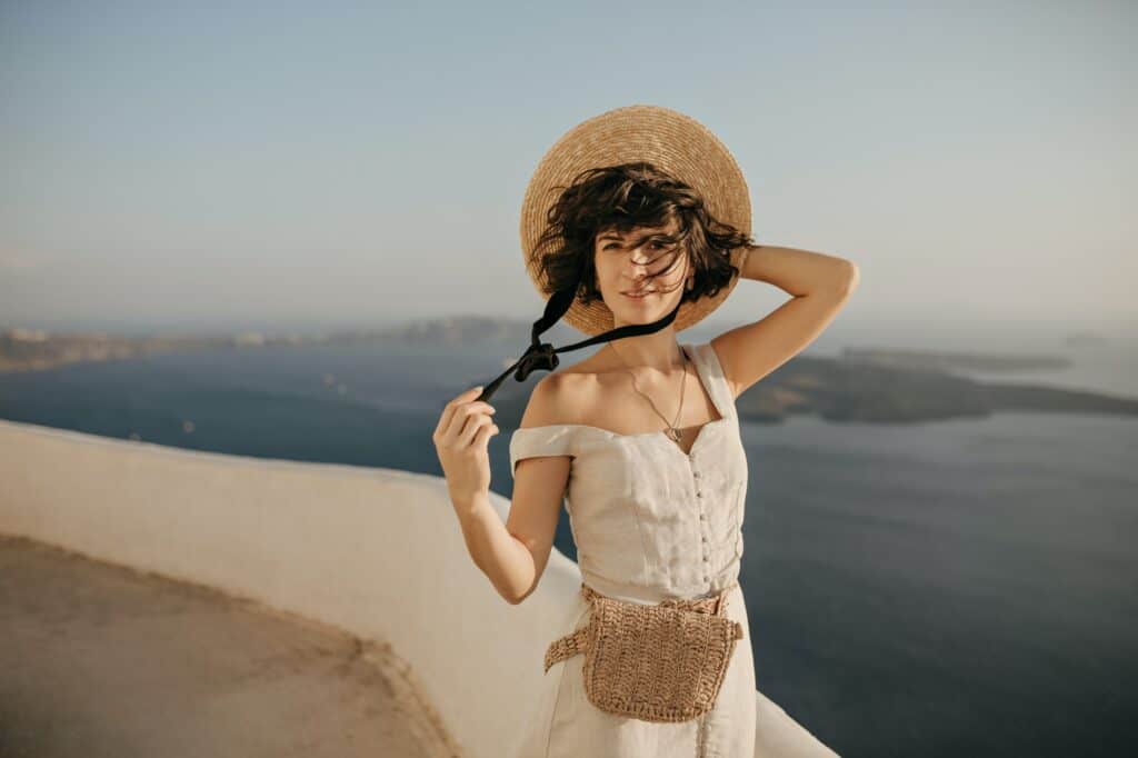 Charming curly brunette woman holds straw hat and smiles. Pretty lady in beige elegant dress poses