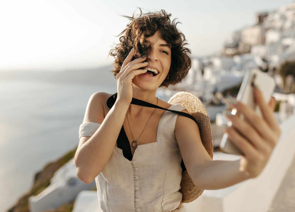 Brunette curly woman in beige dress and straw hat smiles sincerely and takes selfie outside in old